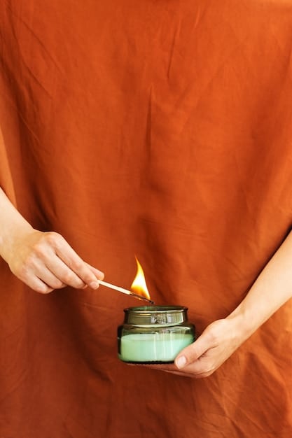 A close-up shot of a person's hands anointing a green candle with essential oil, placed over a work surface scattered with dried herbs and small glass bottles of various essential oils, bathed in warm, natural light.