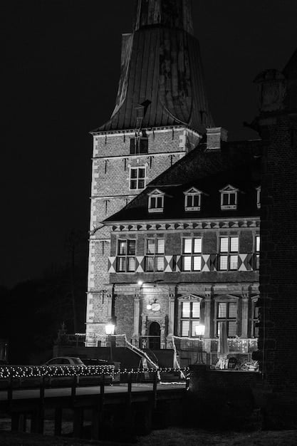 A black and white photo of a historic haunted hotel, with lights on in some of the windows. The setting is dusk, adding to the spooky atmosphere. A road leading up to the hotel is visible in the foreground. It captures the creepy essence of paranormal tourism in the US.