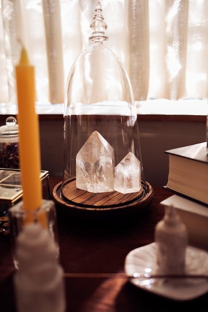 A close-up of a crystal altar featuring a central selenite tower, encircled by various tumbled stones including lapis lazuli, tiger's eye, and carnelian. A small wooden box containing dried herbs sits to the side, and a single white candle flickers gently. The image highlights the diversity of elements that can be included when learning how to create a crystal altar.